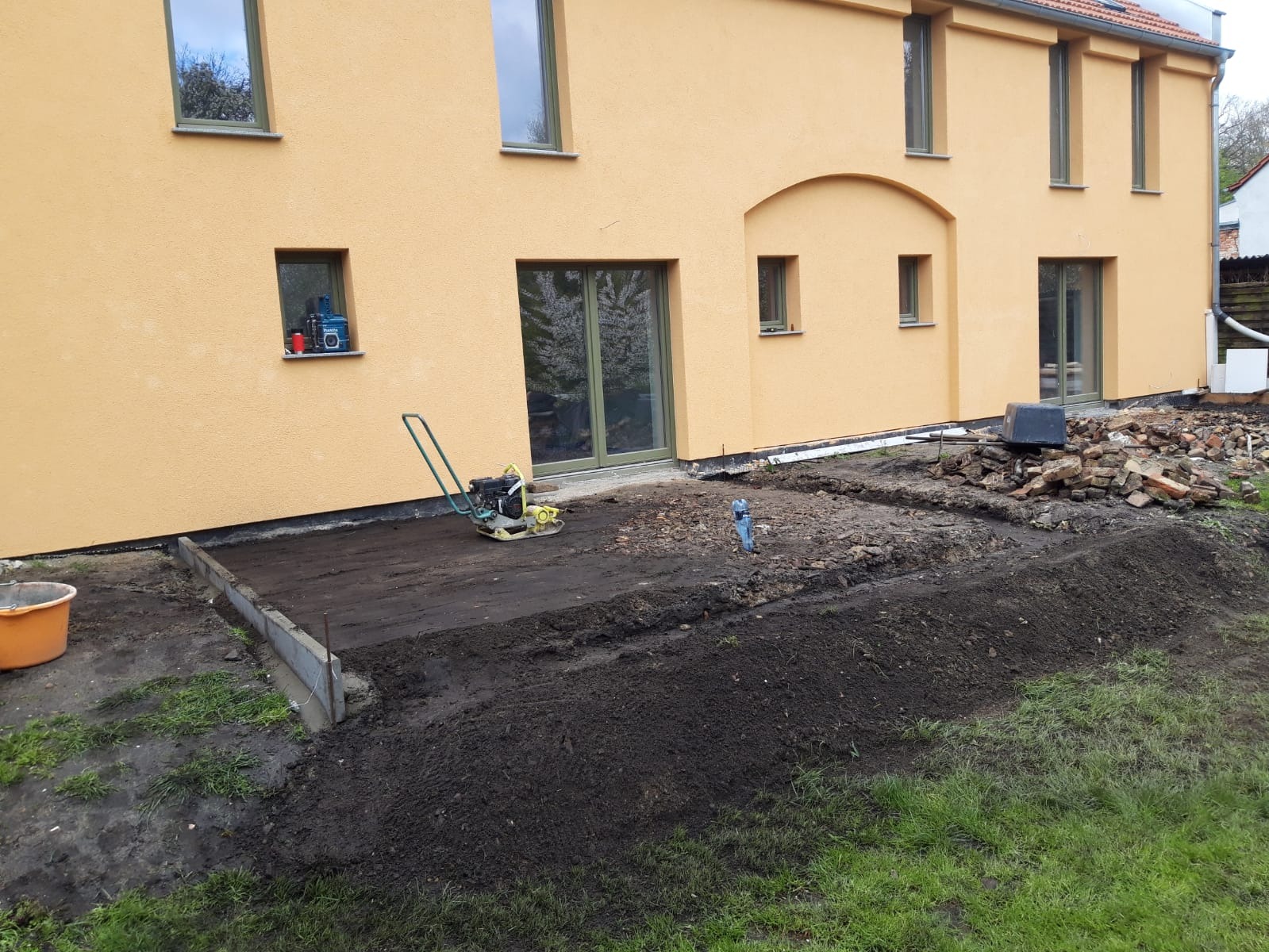 Przygotowanie terenu pod taras przy żółtej elewacji domu, widoczna ubijarka, wykop, obrzeża betonowe i sterta gruzu.