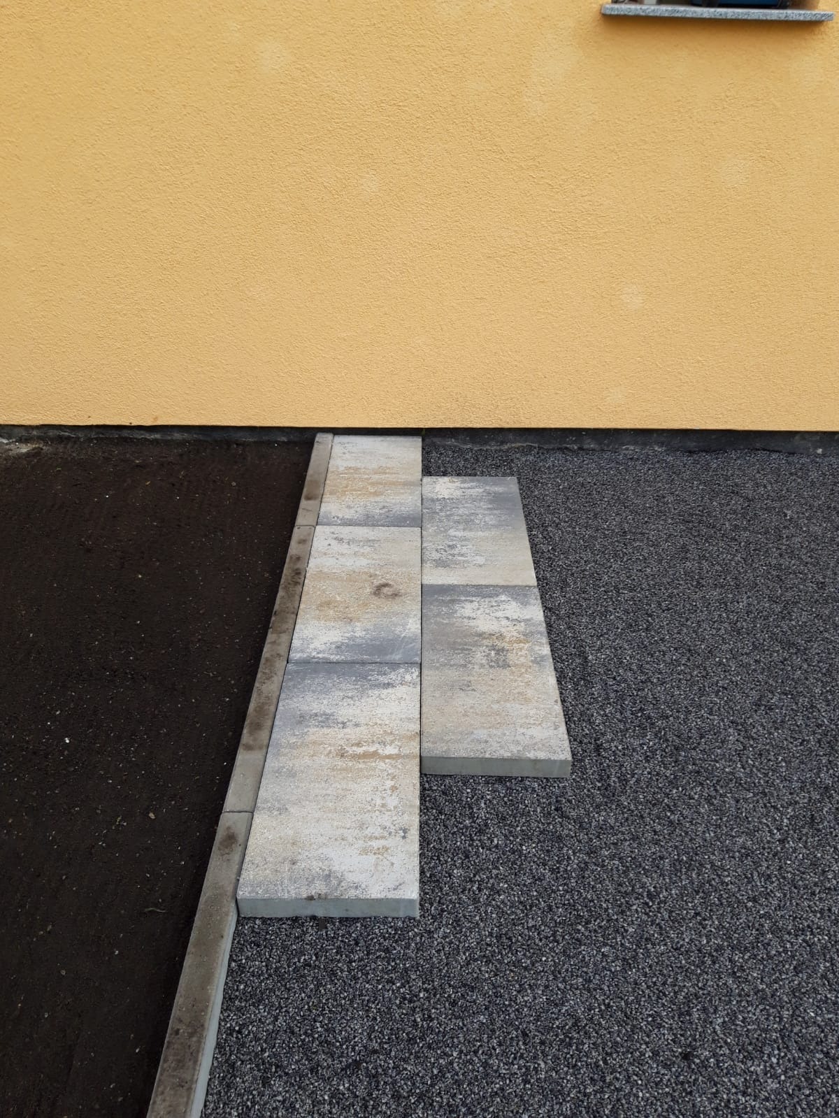 Układanie szarej kostki brukowej przy żółtej ścianie budynku, widoczny fragment okna, czarna ziemia i szary żwir.