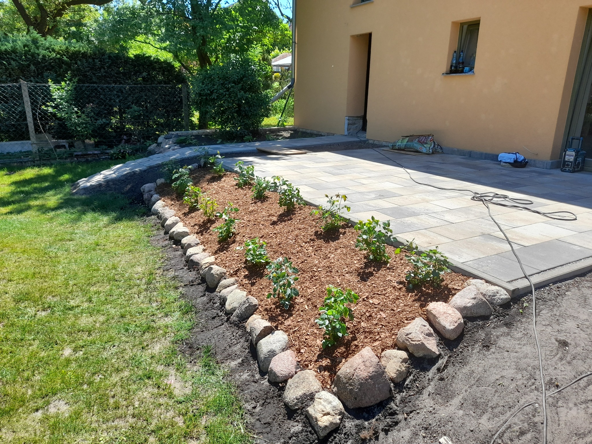 Ułożona kostka brukowa przed budynkiem z nowo zasadzonymi krzewami w korze, obramowanymi kamieniami.