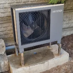 Szara pompa ciepła Neoheat zamontowana na betonowym fundamencie przy ogrodzeniu z betonowych bloczków, z widoczną rurą odprowadzającą i zielonymi krzewami w tle.