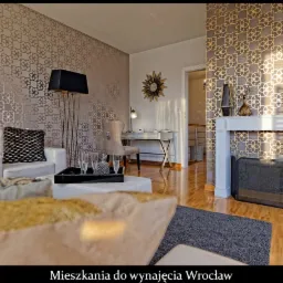 Elegancki salon z wzorzystą tapetą, białym kominkiem, sofą, stolikiem kawowym i drewnianą podłogą, oświetlony popołudniowym słońcem.