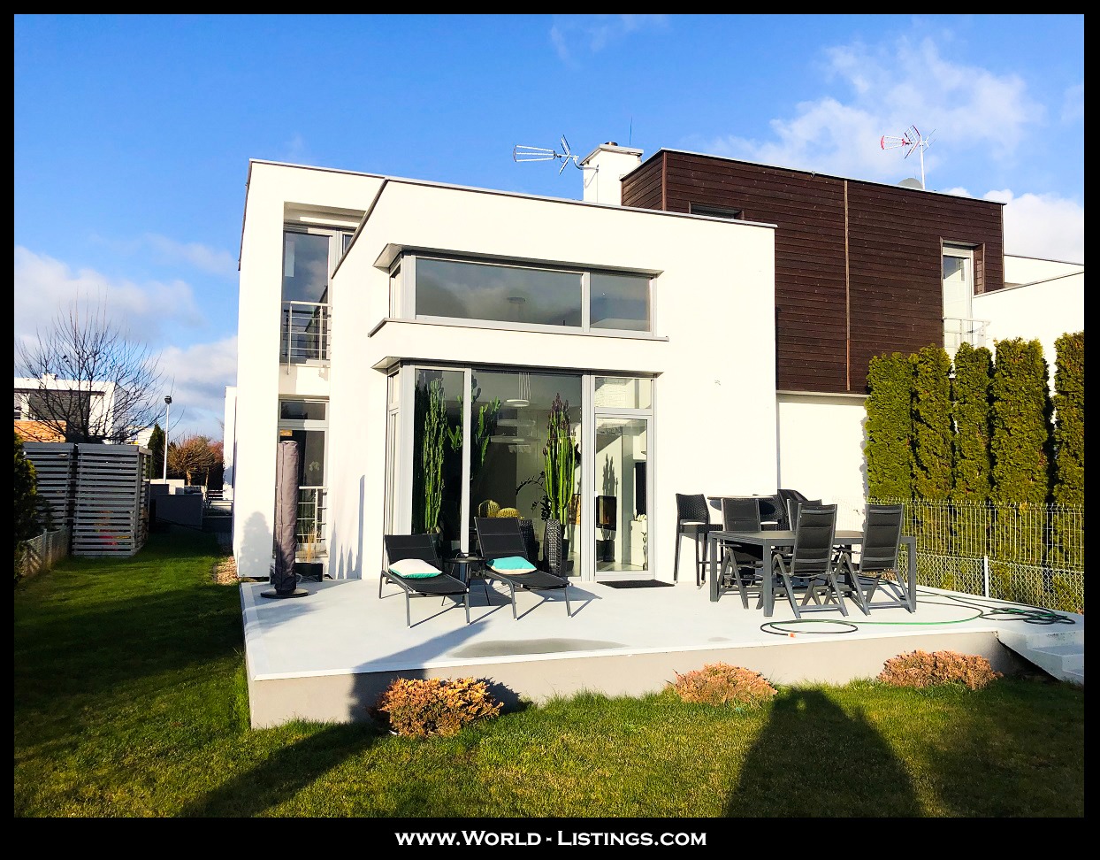 Nowoczesny, dwukondygnacyjny dom z białymi ścianami i ciemnobrązowym drewnianym elementem na górze, z tarasem wyposażonym w czarne leżaki i stół z krzesłami, otoczony zielonym trawnikiem i tujami.