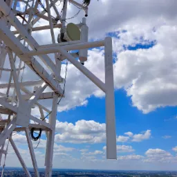 Konstrukcja stalowa wieży telekomunikacyjnej z antenami na tle błękitnego nieba z obłokami i panoramy miasta w oddali.