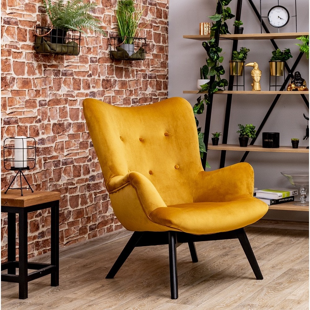 Musztardowy fotel uszak na czarnych nogach, z guzikami na oparciu, na tle ceglanej ściany z roślinami w metalowych koszykach oraz regału z dekoracjami.