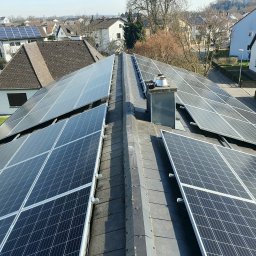 Damian Dembski DecoRoma - Panele fotowoltaiczne na dachu domu jednorodzinnego, widok z góry, komin w tle, osiedle domów w oddali.