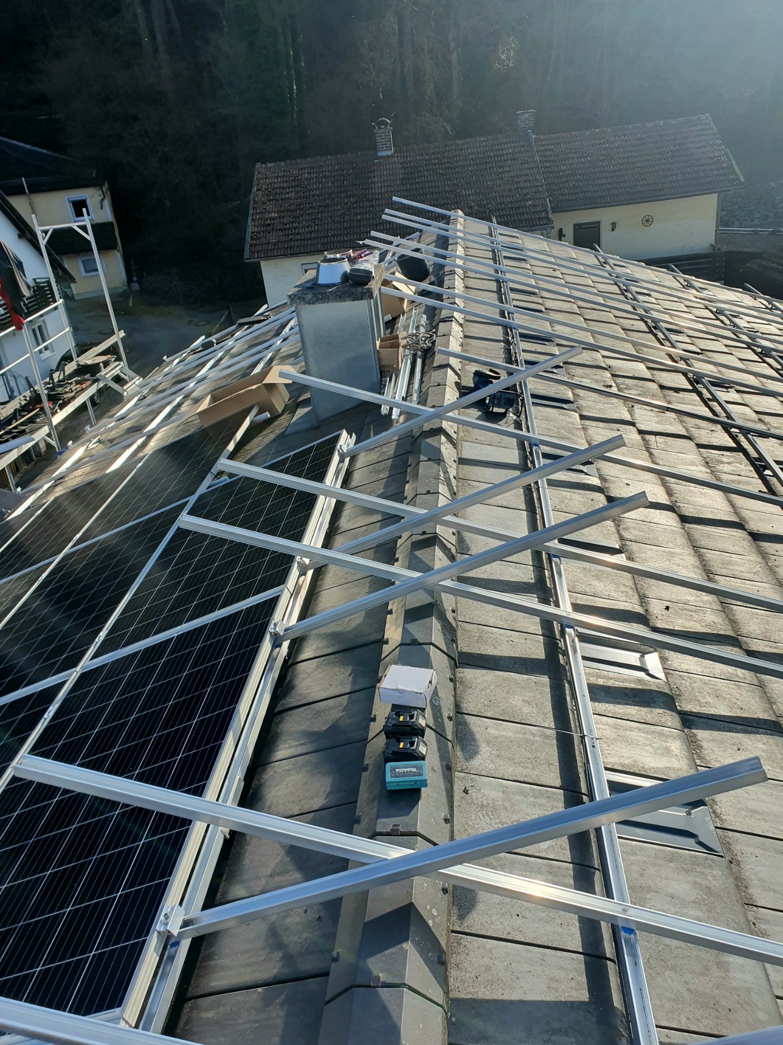 Montaż paneli słonecznych na dachu z szarej dachówki. Widoczne aluminiowe profile konstrukcyjne i częściowo zamontowane panele. W tle budynki mieszkalne i drzewa.