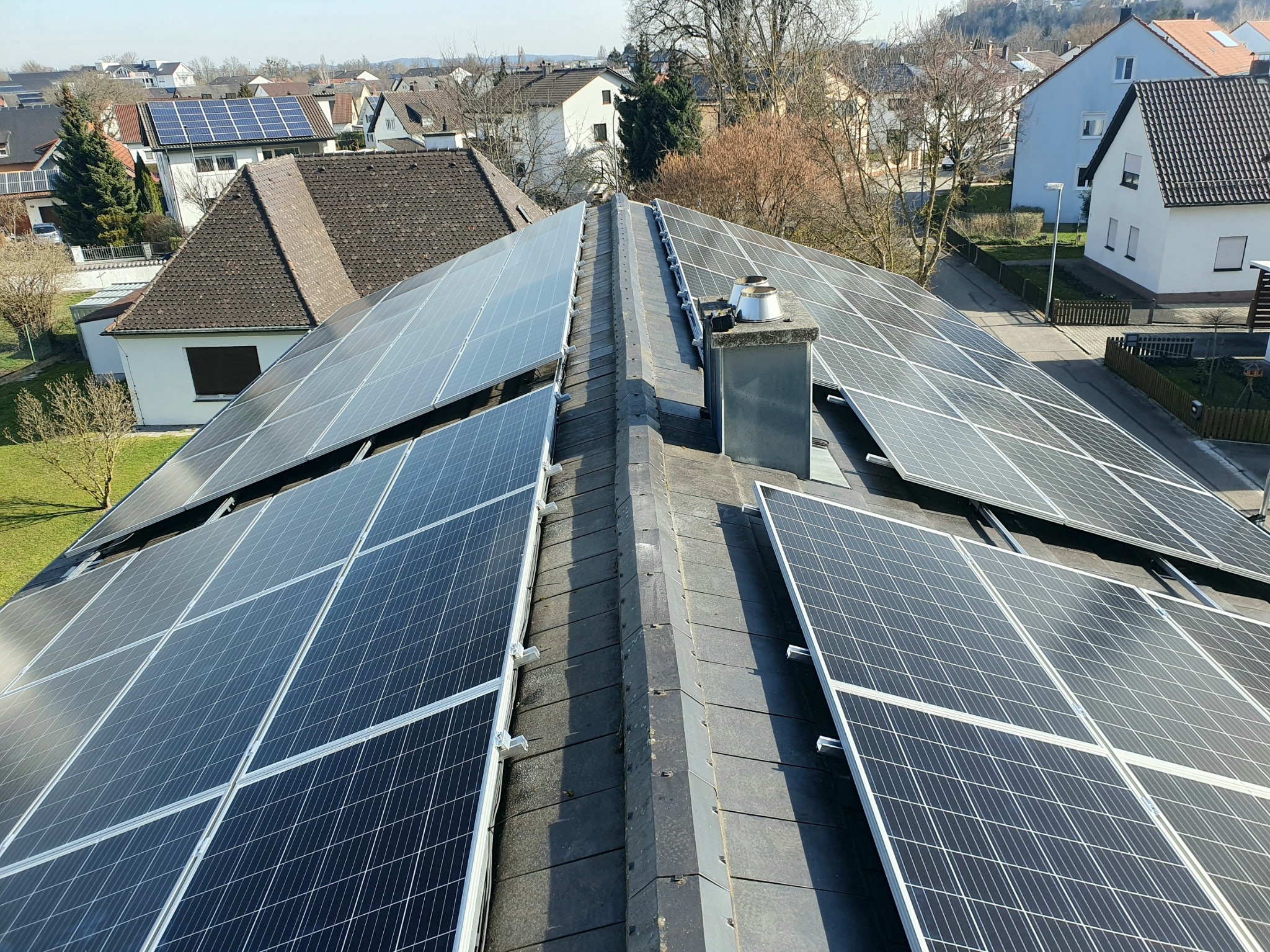 Panele fotowoltaiczne na dachu domu jednorodzinnego, widok z góry, komin w tle, osiedle domów w oddali.