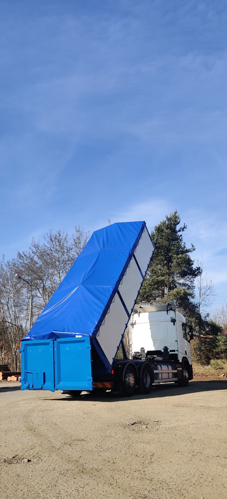 Ciężarówka z otwartą, niebieską plandeką, w trakcie rozładunku kontenera. Widoczne koła, podwozie i fragment kabiny. Tło stanowi niebo i drzewa.