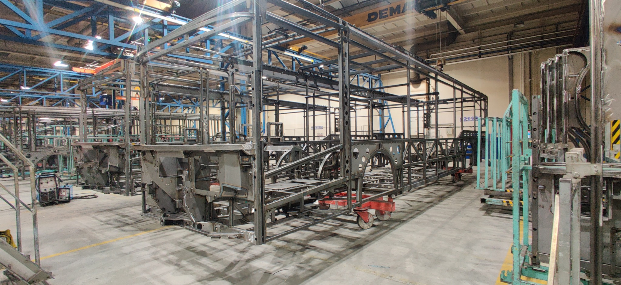 Szkielet autobusu z metalowych profili na hali produkcyjnej, widok z boku, z elementami konstrukcji na wózkach. W tle suwnica DEMAG.