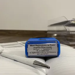 Niebieska pieczątka z danymi 'Biuro Rachunkowe As Taxes' leży na otwartym kalendarzu z długopisem obok, w tle widoczne dokumenty w segregatorze.