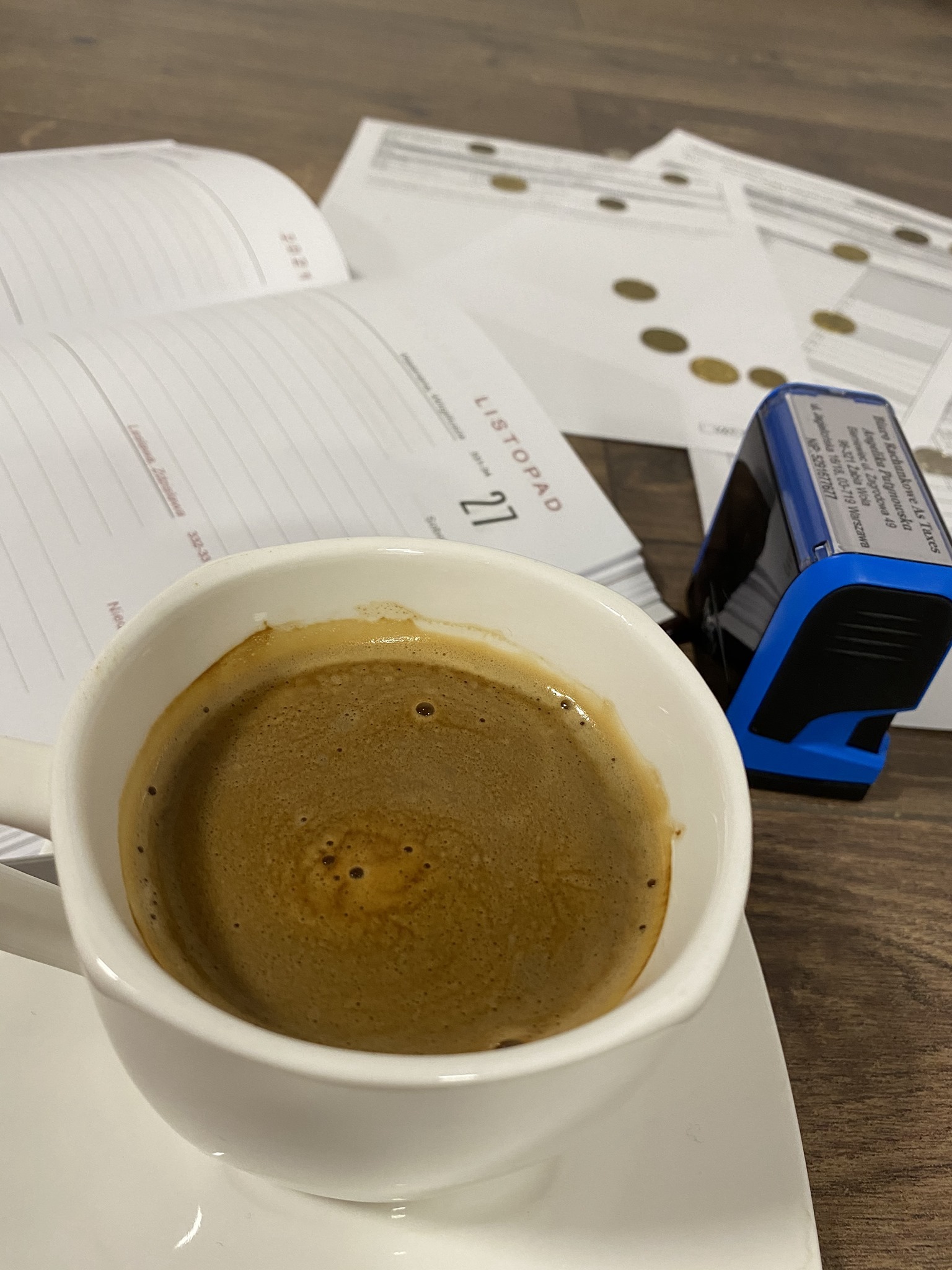 Filiżanka kawy na biurku z otwartym kalendarzem na listopad i stemplowaną pieczątką firmy z danymi adresowymi, sugerująca przerwę w pracy.