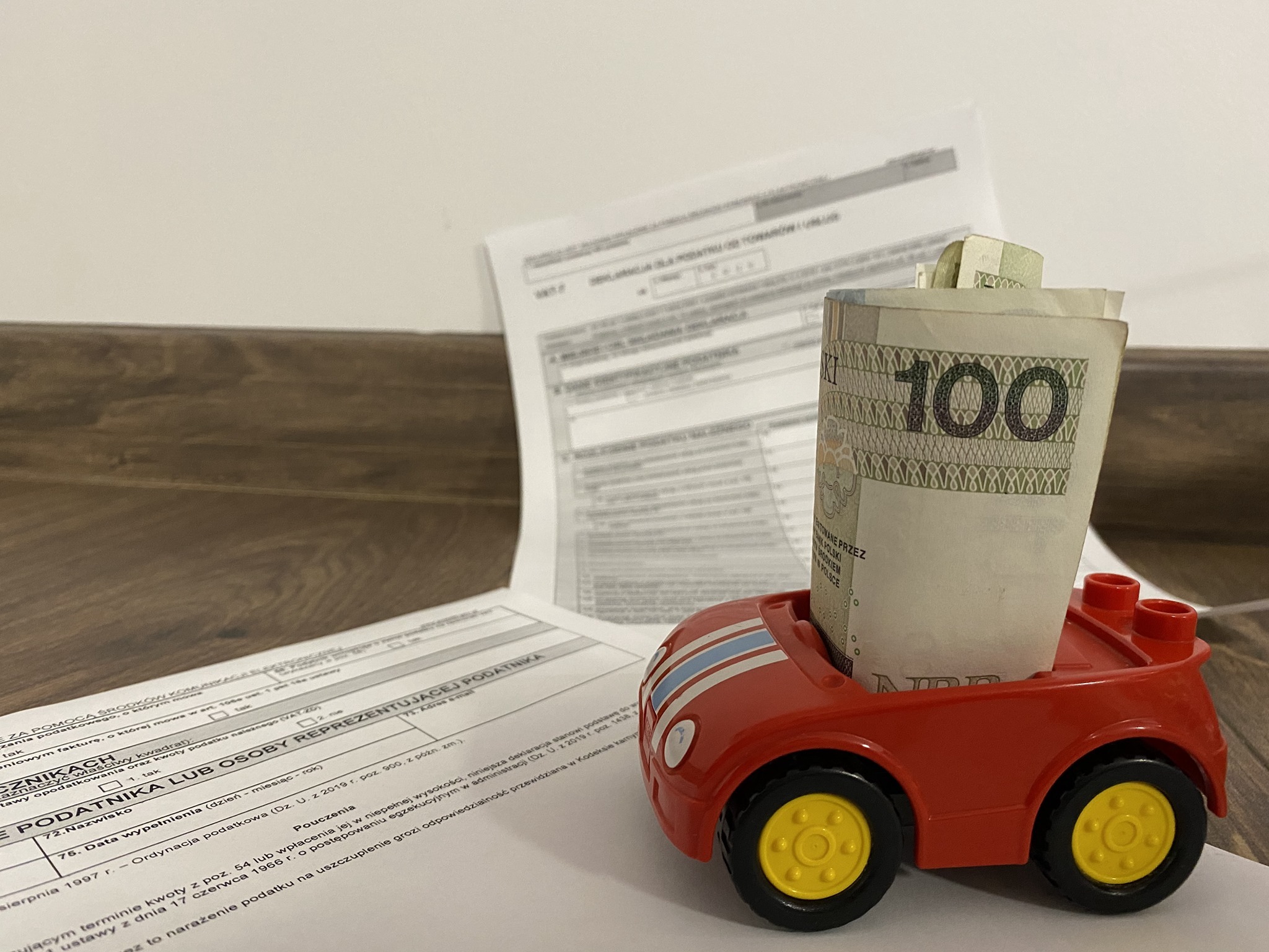 Formularz podatkowy z zabawką w kształcie czerwonego samochodu wypełnionego zwitkiem banknotów 100 zł, symbolizujący koszty związane z podatkami.