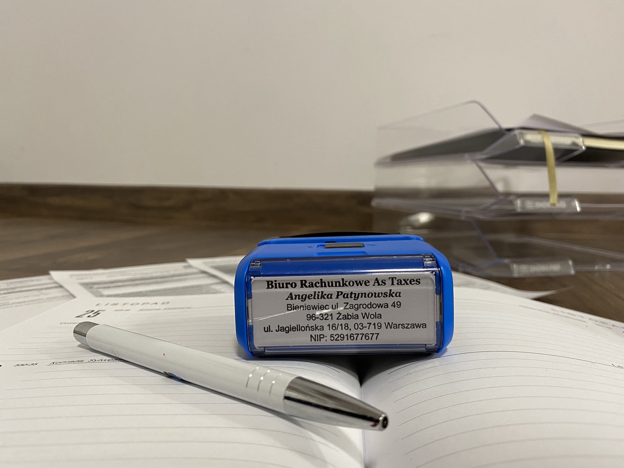 Niebieska pieczątka z danymi 'Biuro Rachunkowe As Taxes' leży na otwartym kalendarzu z długopisem obok, w tle widoczne dokumenty w segregatorze.
