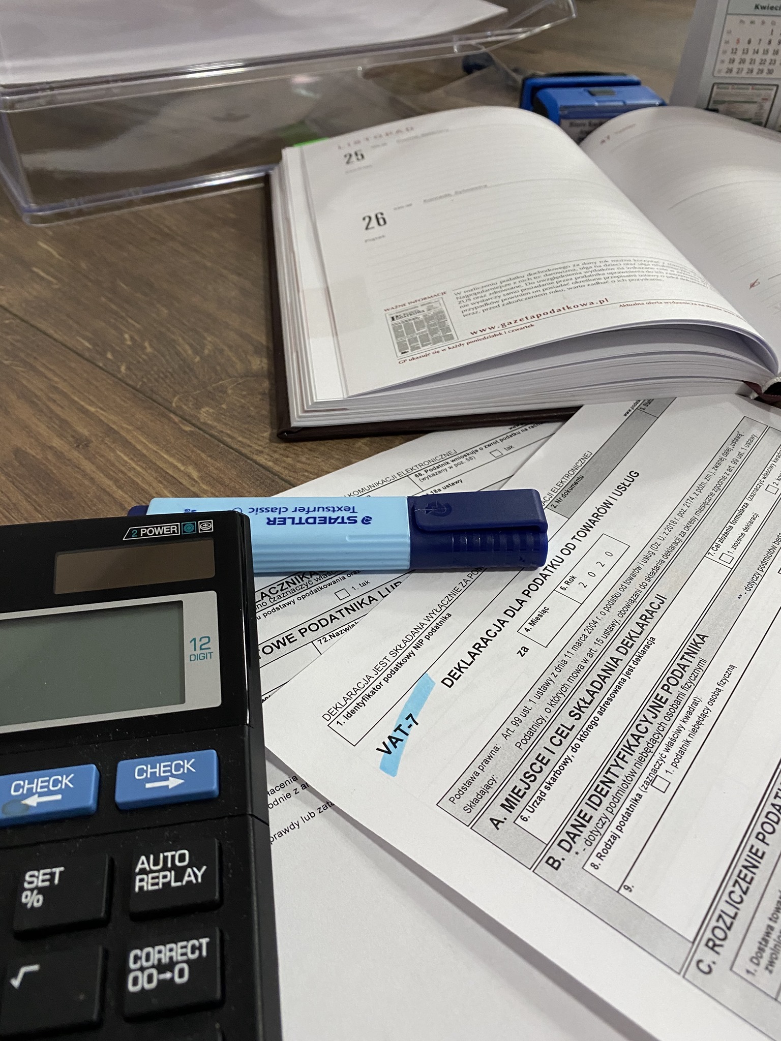 Formularz VAT-7, kalkulator, otwarty kalendarz i zakreślacz na biurku, przygotowane do rozliczeń podatkowych.