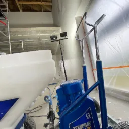 Wnętrze w trakcie prac w Gorzyce Wielkie, widoczny agregat malarski TexSpray T-Max 506, przygotowany do natrysku na ścianach zabezpieczonych folią, z lampami oświetlającymi sufit.