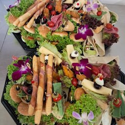 Strefa Polskiego Smaku - Dwa czarne, plastikowe półmiski z różnorodnymi przekąskami: mini-burgery, tortille, grissini owinięte szynką, sery, sałatki, koreczki, udekorowane kwiatami orchidei i listkami sałaty.