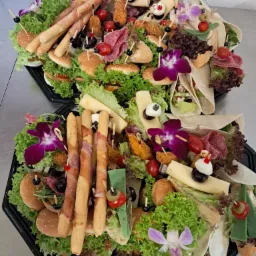 Dwa czarne, plastikowe półmiski z różnorodnymi przekąskami: mini-burgery, tortille, grissini owinięte szynką, sery, sałatki, koreczki, udekorowane kwiatami orchidei i listkami sałaty.