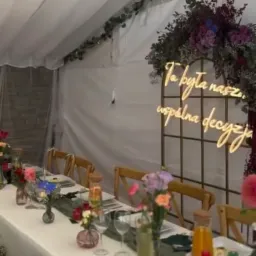 Uroczysty stół w namiocie, dekoracje kwiatowe, neonowy napis 'To była nasza wspólna decyzja'. Przygotowania do wesela, detale wystroju.