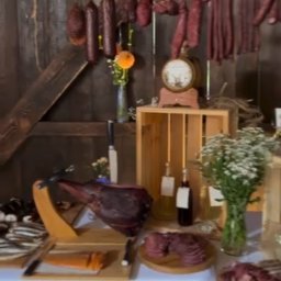 Strefa Polskiego Smaku - Rustykalny stół z wędlinami, szynką na stojaku, kwiatami w wazonie i dekoracjami na tle drewnianej ściany. Ujęcie z imprezy w stylu wiejskim.