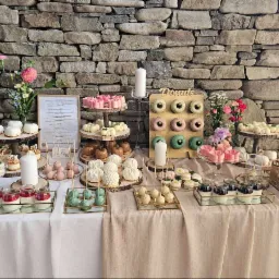 Bogato zastawiony stół z deserami: donuty, ciasta, desery w słoiczkach. Kamienne tło, świeże kwiaty i świece dodają elegancji. Idealne na przyjęcie.