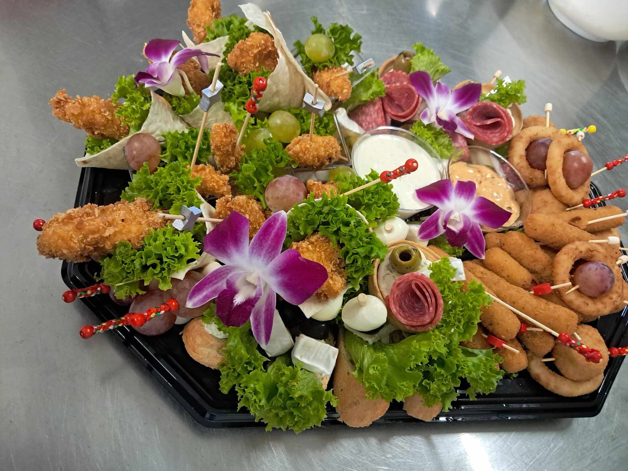 Dekoracyjna taca z przekąskami: smażone kąski na patyku z dekoracyjnymi zakończeniami, mini tortille, winogrona, salami, ser mozzarella, oliwki i sałata, ozdobiona fioletowymi kwiatami orchidei.