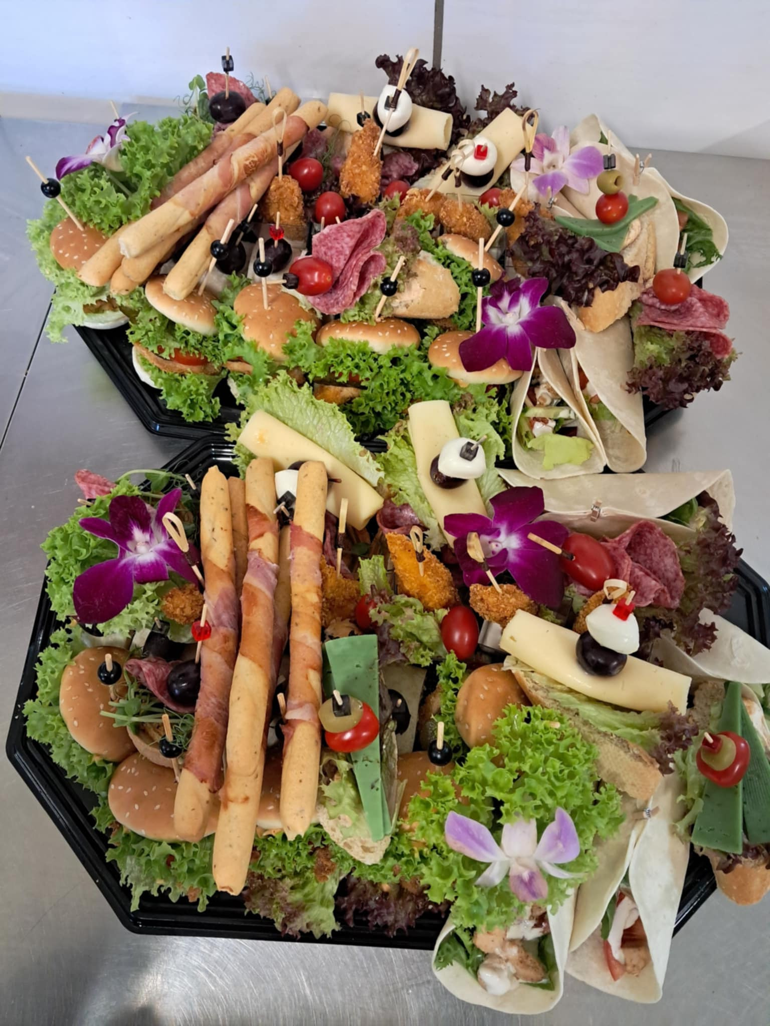 Dwa czarne, plastikowe półmiski z różnorodnymi przekąskami: mini-burgery, tortille, grissini owinięte szynką, sery, sałatki, koreczki, udekorowane kwiatami orchidei i listkami sałaty.