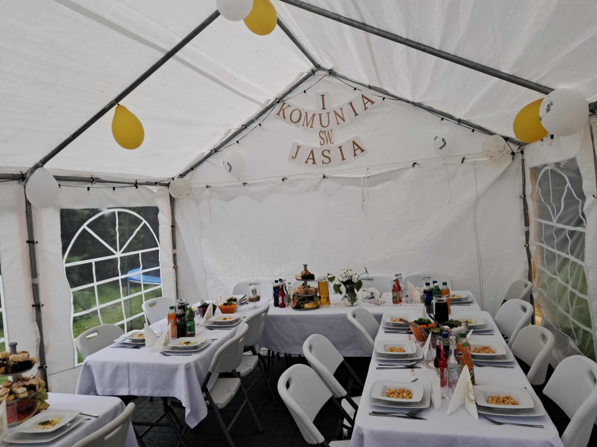 Uroczystość Pierwszej Komunii Świętej Jasia w białym namiocie, stoły z białymi obrusami, białe krzesła, balony białe i żółte, dekoracja z napisem 'I Komunia Św. Jasia'.