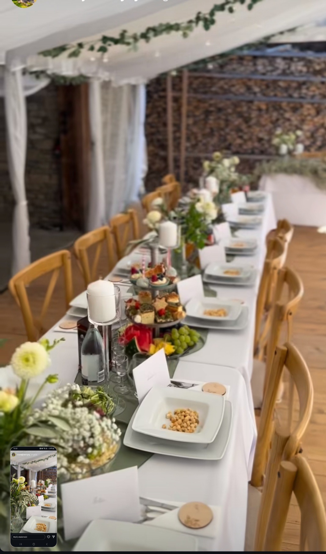 Uroczysty stół w namiocie: biały obrus, kwiatowe dekoracje, świece, elegancka zastawa. Widoczne drewniane krzesła i elementy dekoracyjne z zieleni.