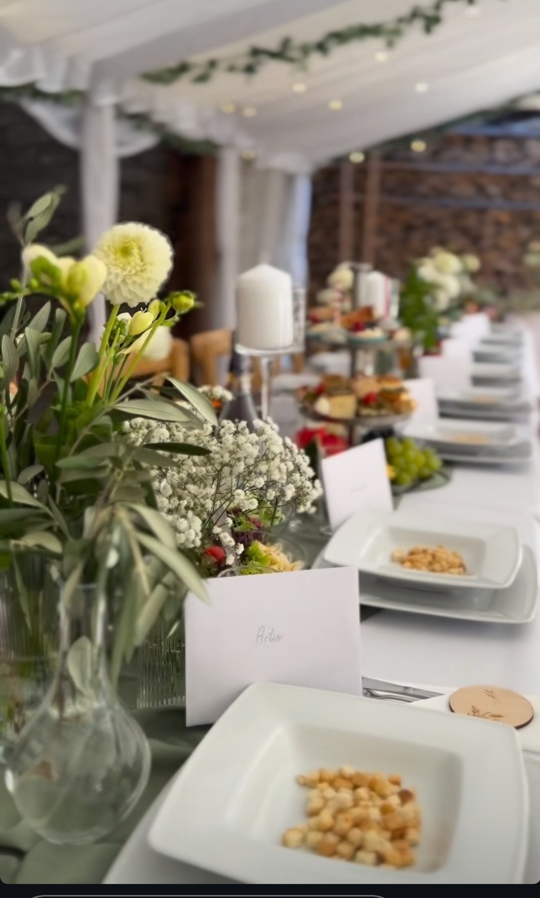 Uroczysty stół cateringowy pod namiotem: białe obrusy, kwiatowe dekoracje, elegancka zastawa i imienne winietki, tworzące wyrafinowaną oprawę eventu.