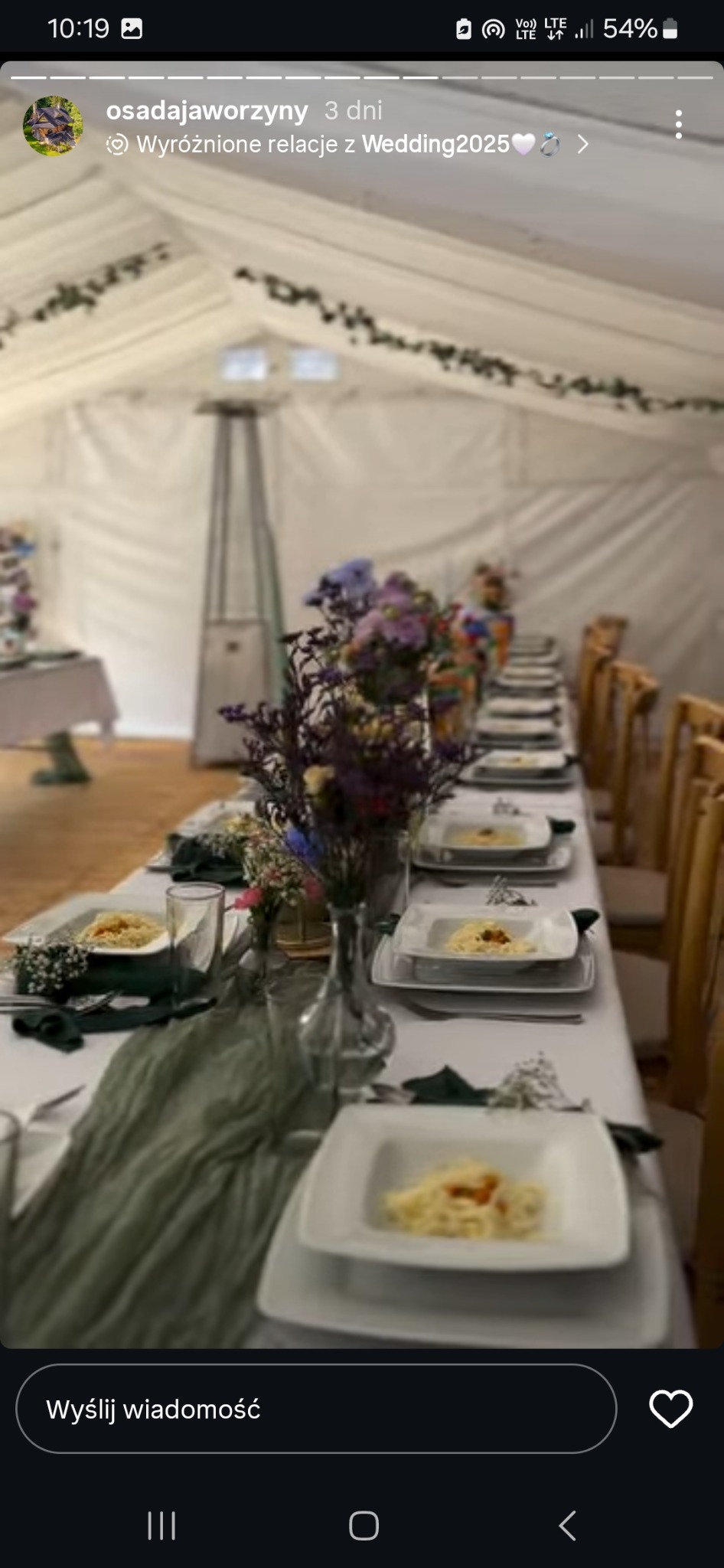 Udekorowany, długi stół z białym obrusem, kwiatami i zastawą w namiocie. Danie na talerzach, zielony bieżnik. Przygotowania do przyjęcia weselnego.