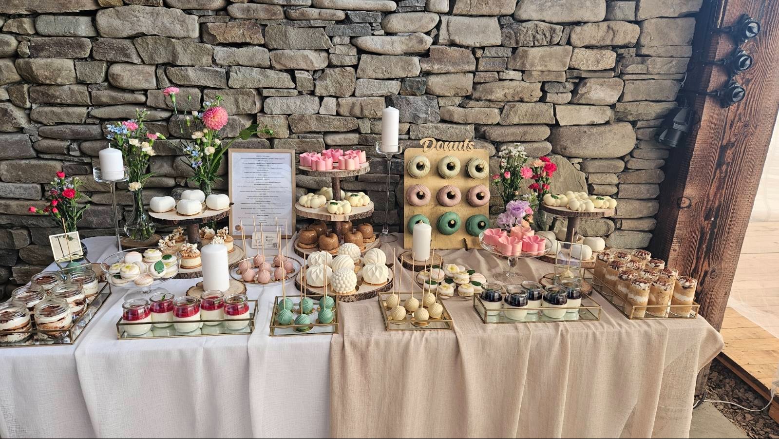 Bogato zastawiony stół z deserami: donuty, ciasta, desery w słoiczkach. Kamienne tło, świeże kwiaty i świece dodają elegancji. Idealne na przyjęcie.