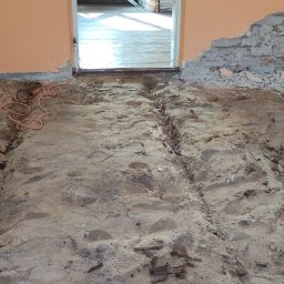 PHU Adam Karpiński - Pomieszczenie w trakcie demontażu podłogi: widoczne ślady po usuniętych deskach, gruz na ścianie i narzędzia elektryczne.