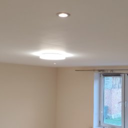 PHU Adam Karpiński - Jasne wnętrze z kremowymi ścianami, białym sufitem z trzema punktowymi lampami i jedną okrągłą lampą sufitową, okno z widokiem na budynek z czerwonej cegły.