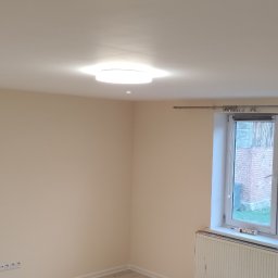 PHU Adam Karpiński - Jasny pokój po remoncie z kremowymi ścianami, oświetleniem punktowym w suficie i okrągłą lampą sufitową. Widoczne okno z widokiem na budynek gospodarczy i grzejnik pod oknem.