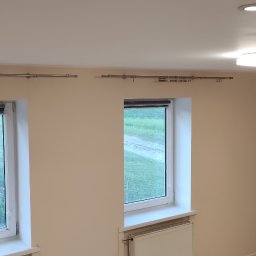 PHU Adam Karpiński - Jasne wnętrze po remoncie z dwoma oknami z roletami, grzejnikiem pod jednym z nich, karniszami i oświetleniem sufitowym.