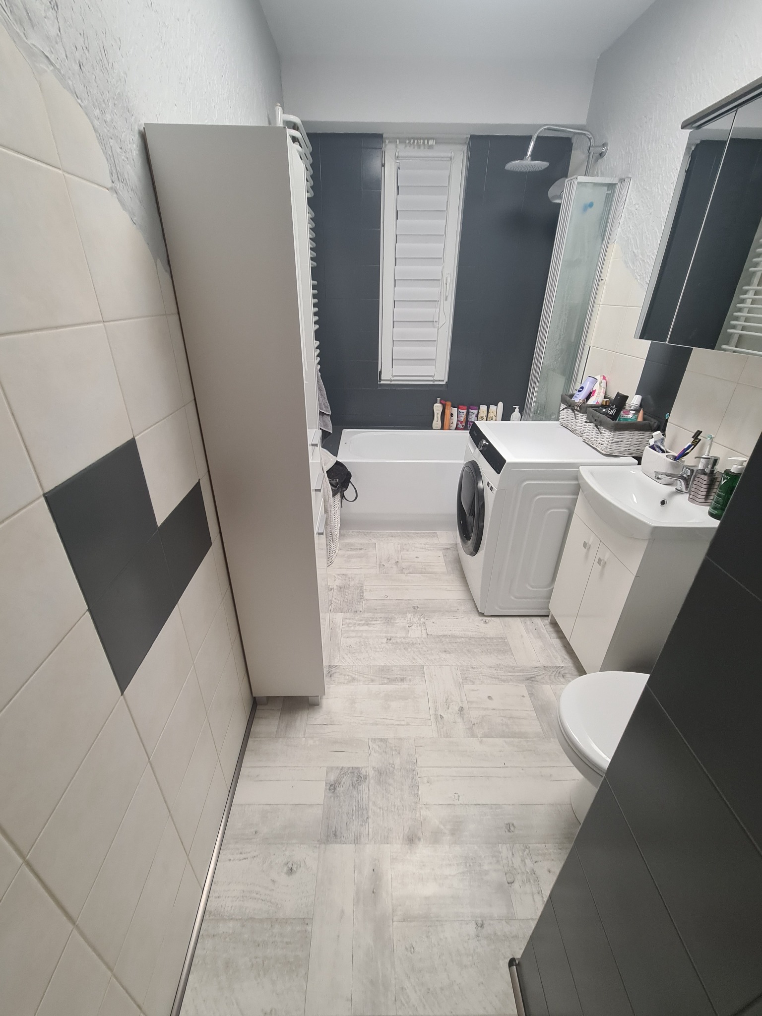 Wnętrze wyremontowanej łazienki z pralką, umywalką i kabiną prysznicową, wykończonej w odcieniach bieli i szarości z akcentami czerni na ścianach i podłodze.