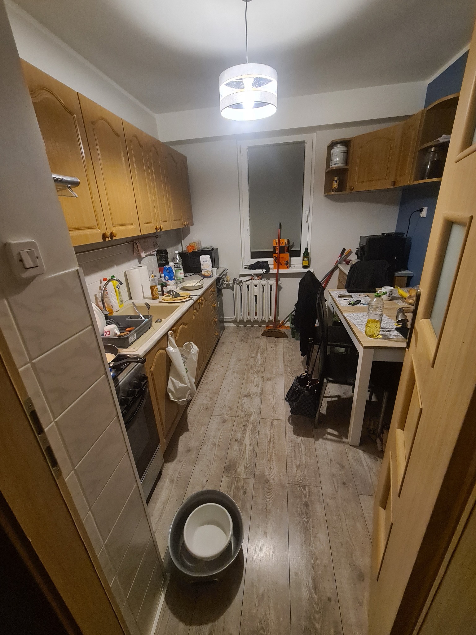 Widok na wnętrze kuchni z drewnianymi szafkami, stołem, grzejnikiem i podłogą wyłożoną panelami imitującymi drewno. W zlewie naczynia, na stole przedmioty codziennego użytku. Uwagę zwraca wiadro...