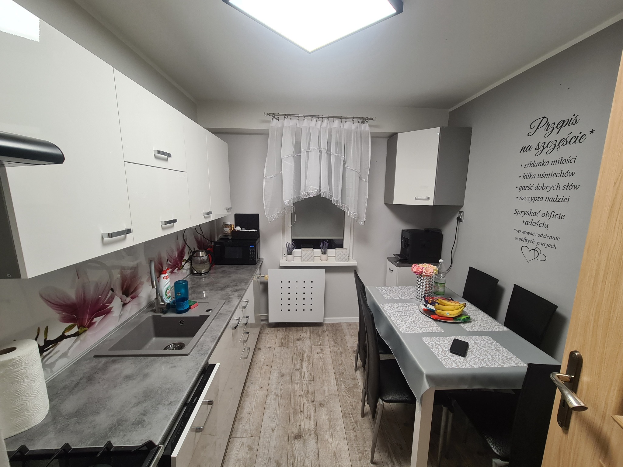 Nowoczesna kuchnia z białymi szafkami, szarym blatem z motywem kwiatowym, drewnopodobną podłogą i tekstem 'Przepis na szczęście' na ścianie.