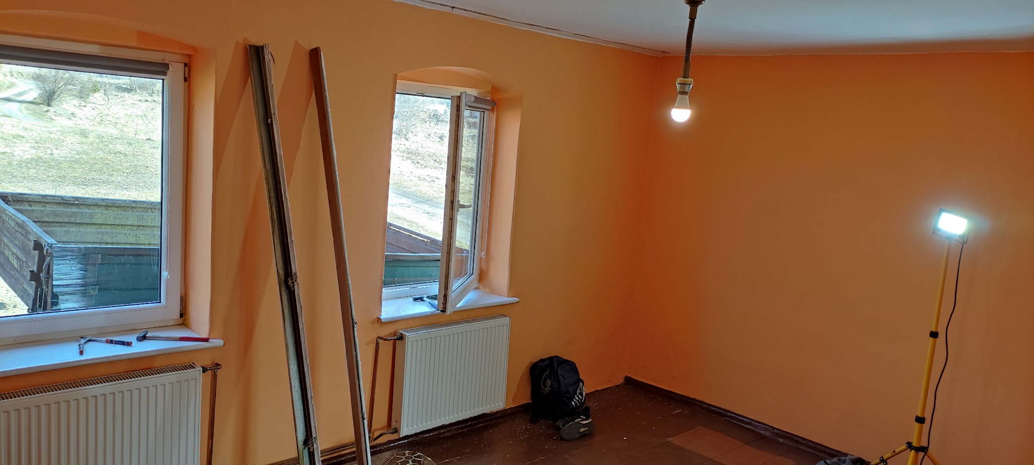 Pomarańczowy pokój w trakcie prac, widoczne dwa okna, jedno otwarte, ramy okienne oparte o ścianę, narzędzia na parapecie, grzejnik, lampa pod sufitem i reflektor na statywie.