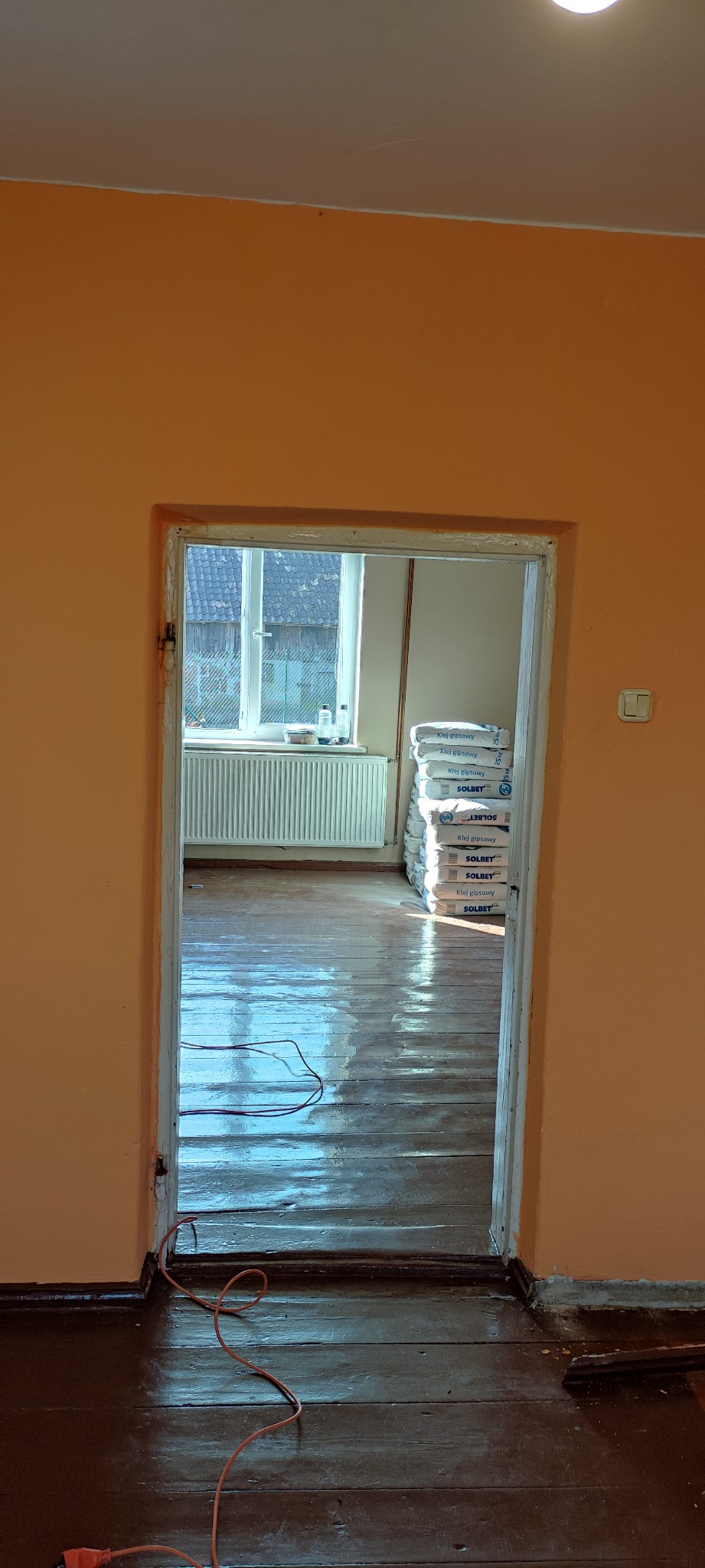 Pokój w trakcie remontu z widoczną stertą worków z klejem gipsowym Solbet w tle, świeżo polakierowana podłoga parkietowa odbija światło, pomarańczowe ściany i widok na okno zza futryny drzwi.