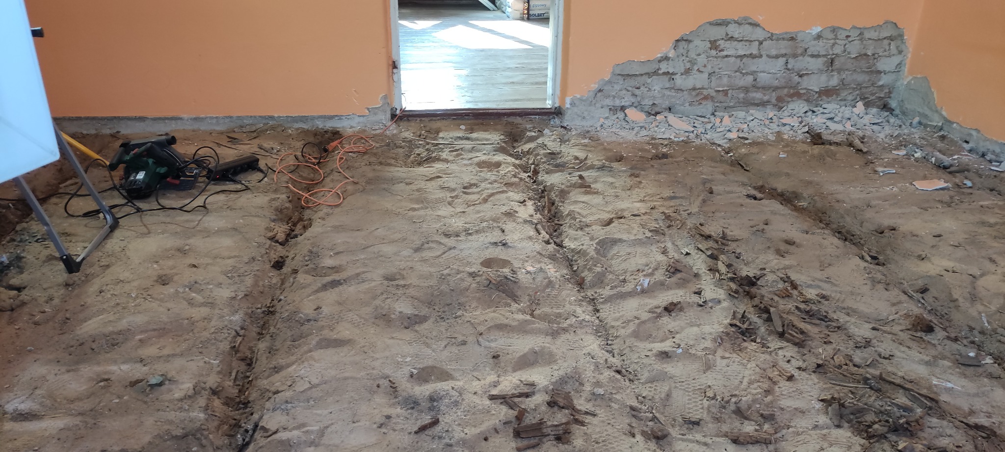 Pomieszczenie w trakcie demontażu podłogi: widoczne ślady po usuniętych deskach, gruz na ścianie i narzędzia elektryczne.