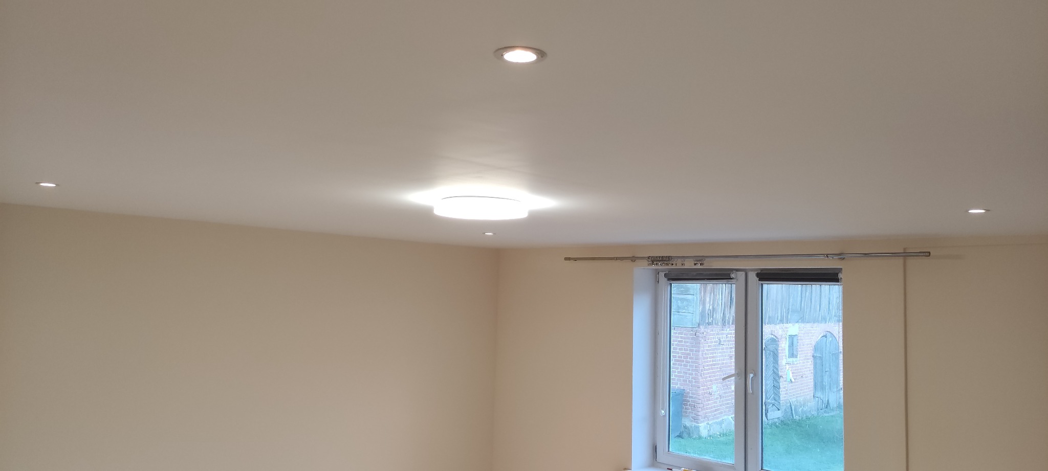 Jasne wnętrze z kremowymi ścianami, białym sufitem z trzema punktowymi lampami i jedną okrągłą lampą sufitową, okno z widokiem na budynek z czerwonej cegły.