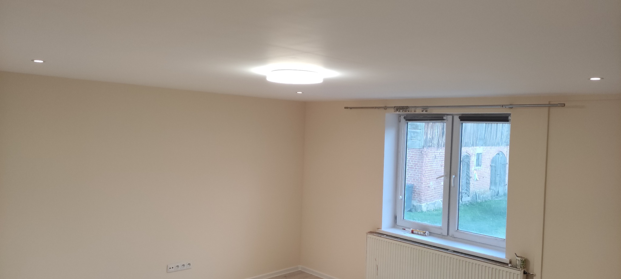 Jasny pokój po remoncie z kremowymi ścianami, oświetleniem punktowym w suficie i okrągłą lampą sufitową. Widoczne okno z widokiem na budynek gospodarczy i grzejnik pod oknem.