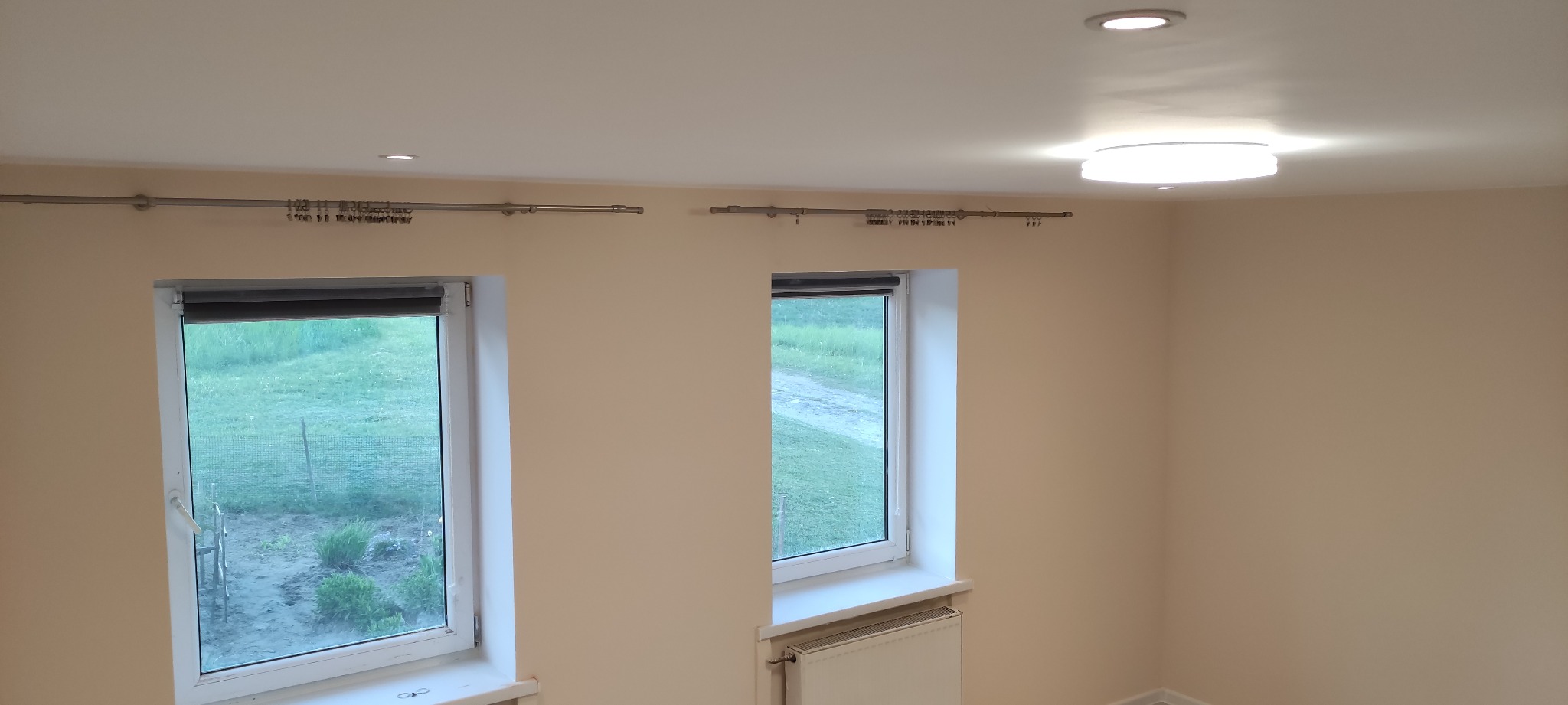 Jasne wnętrze po remoncie z dwoma oknami z roletami, grzejnikiem pod jednym z nich, karniszami i oświetleniem sufitowym.