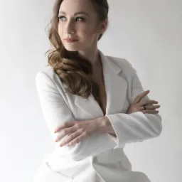 Portret kobiety w białym garniturze, z rękami skrzyżowanymi na piersiach, patrzącej w lewo, na jasnym tle.