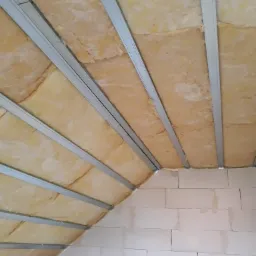 Żółta wełna mineralna izolująca skośny sufit poddasza, zamocowana między metalowymi profilami, widoczne bloczki betonowe ściany.