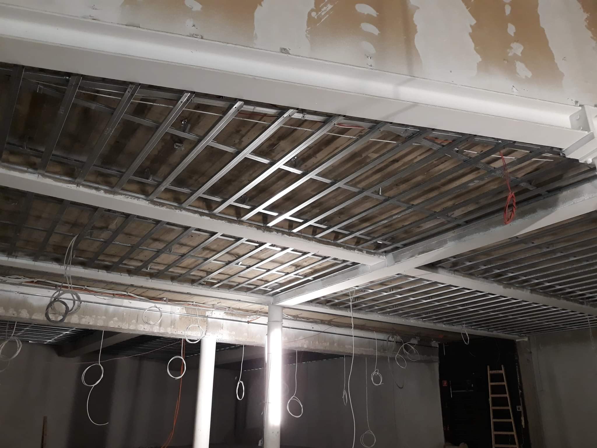 Stelaż sufitowy z metalowych profili, widoczne kable elektryczne, ściany w stanie surowym przed wykończeniem.