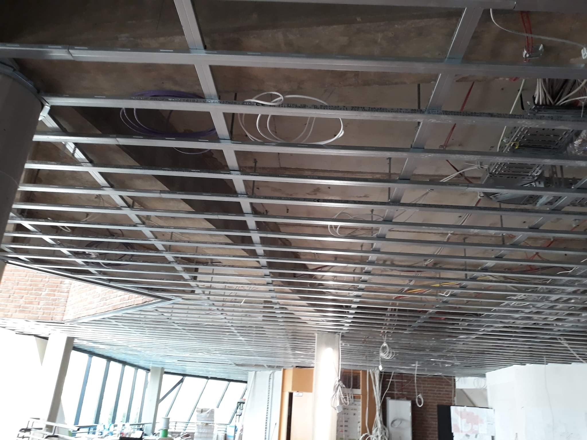 Metalowa konstrukcja sufitowa w trakcie montażu, widoczne przewody elektryczne i rury wentylacyjne, surowy betonowy strop powyżej, duże okna w tle.