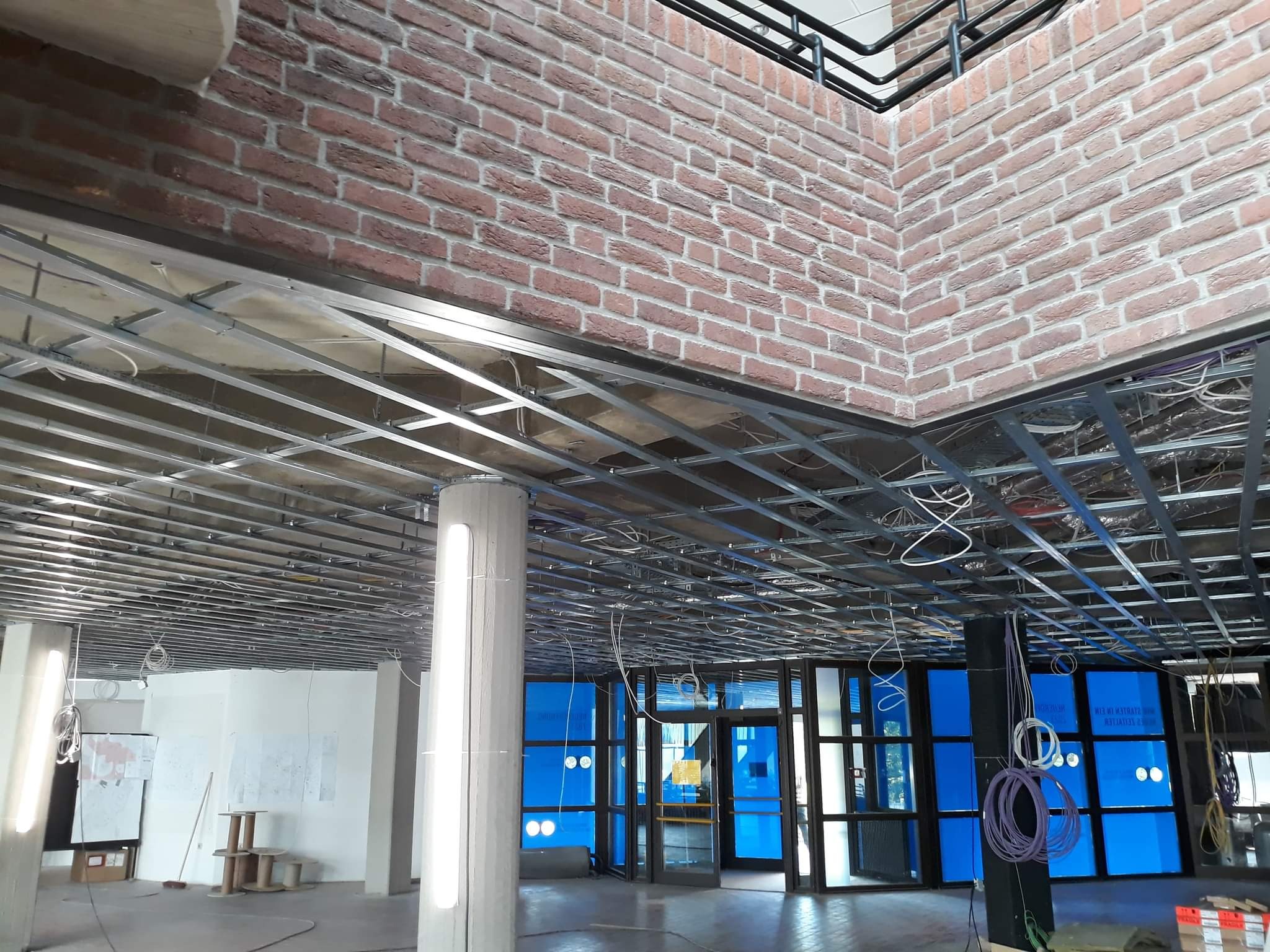 Metalowa konstrukcja sufitu podwieszanego w trakcie montażu, odsłonięta instalacja elektryczna, fragment ściany z cegły dekoracyjnej, widoczne kolumny i przeszklenia.