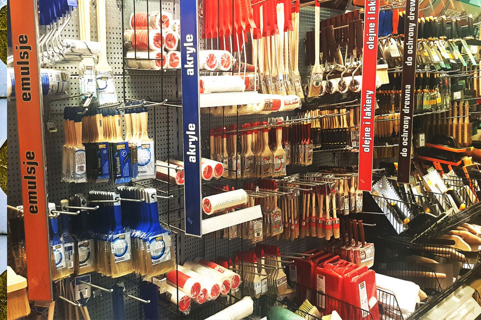 Różnorodność pędzli, wałków i akcesoriów malarskich na metalowym regale w sklepie budowlanym, z etykietami wskazującymi na emulsje, akryle oraz olejne lakiery i preparaty do ochrony drewna.